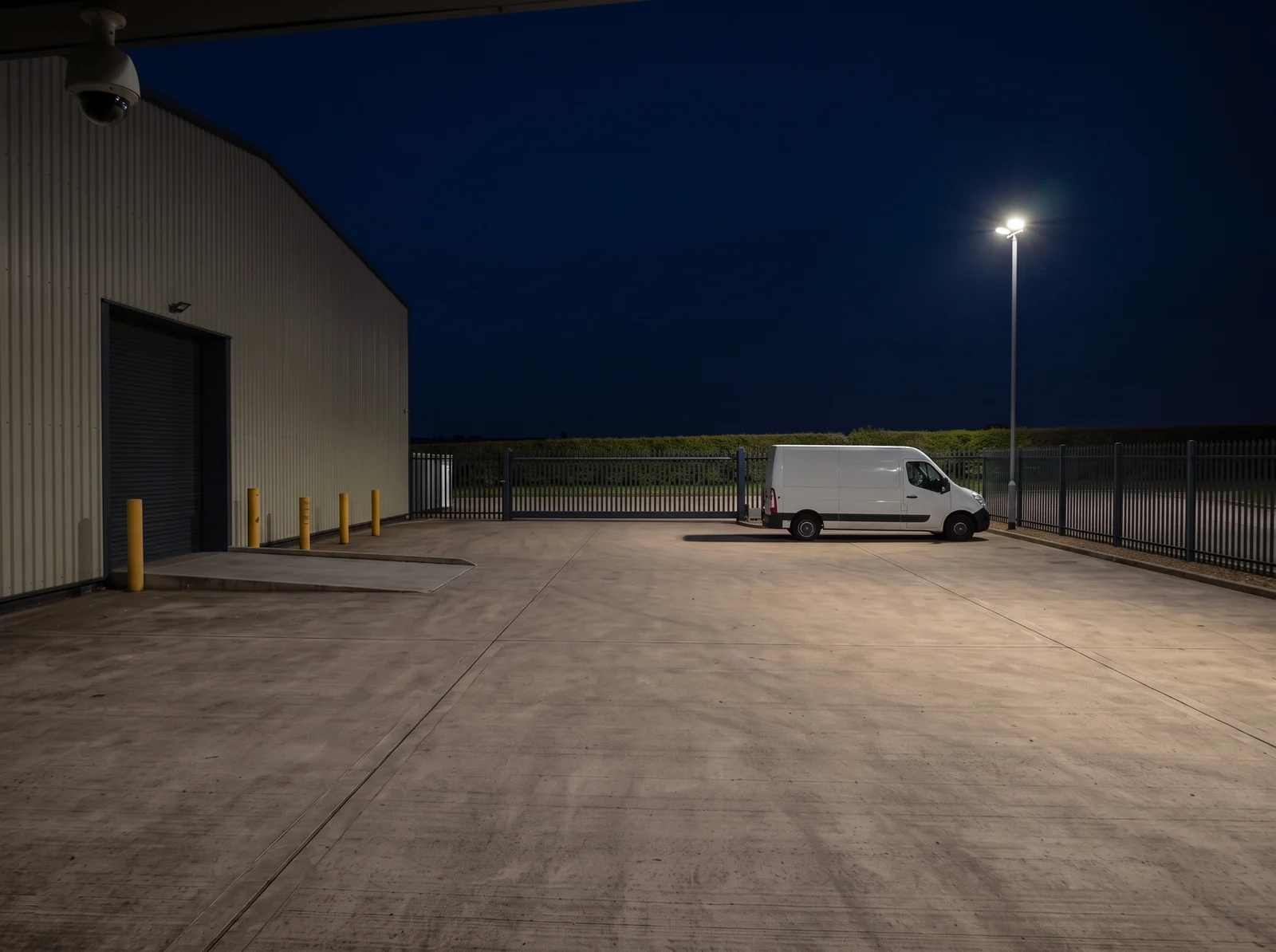 Derselbe Betriebshof bei Nacht — Fahrzeugfarbe, Poller und Hecke bleiben unterscheidbar